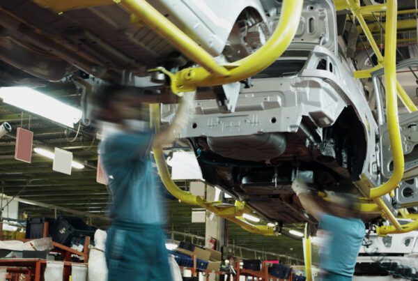 Two workers wearing green shirts and blue pants work underneath a car in an automobile factory. There are assembly lines, partially constructed cars, tools and electrical equipment in the background.