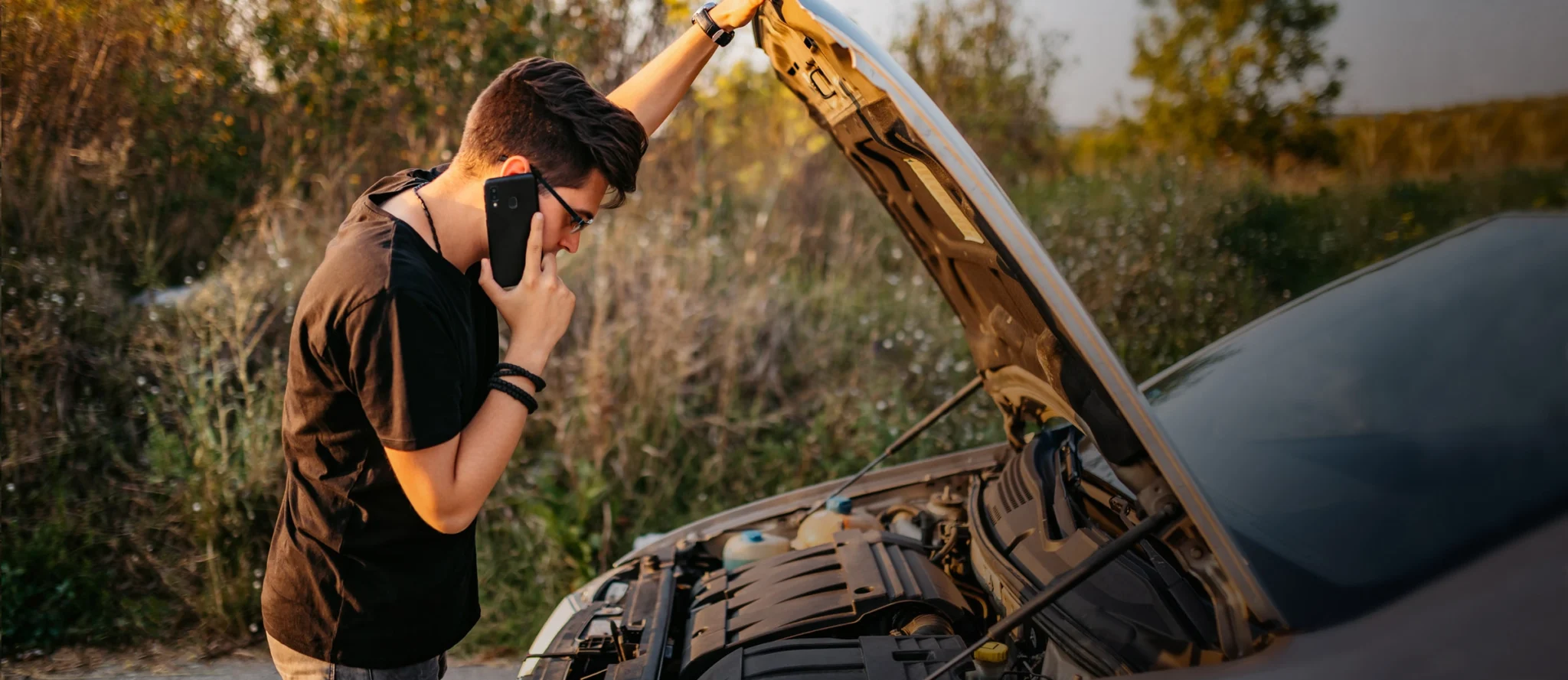 Guy with his hood popped open on the side of the road with his car broken down. He is on the phone looking into his engine.